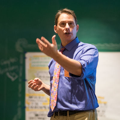 Professor Mark Salzer stands at the front of a classroom with his left hand outstretched while delivering a lecture