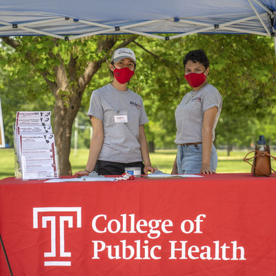 Two students wearing masks stand behind a table topped with a red tablecloth with a white College of Public Health logo on it