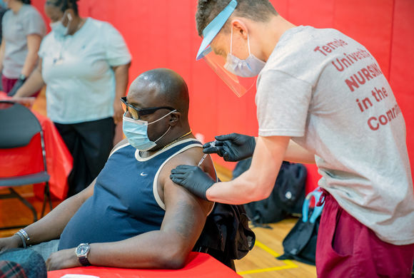 a nursing student administers a covid-19 vaccine into the arm of a seated man in a gymnasium.