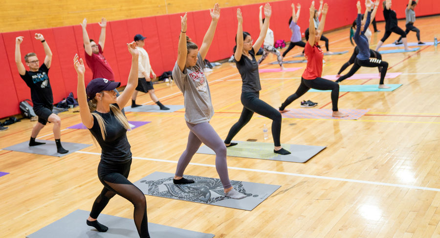 Students in a gym standing on mats with their arms raised doing yoga