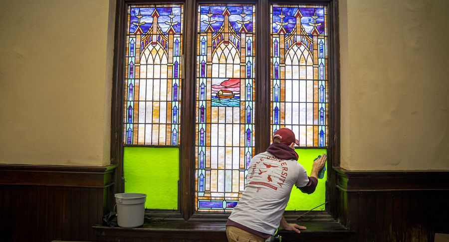 A man holding a rag wipes down a stained glass window inside a church