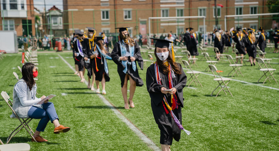 Students in caps and gowns stand and walk to the graduation stage on geasey field 
