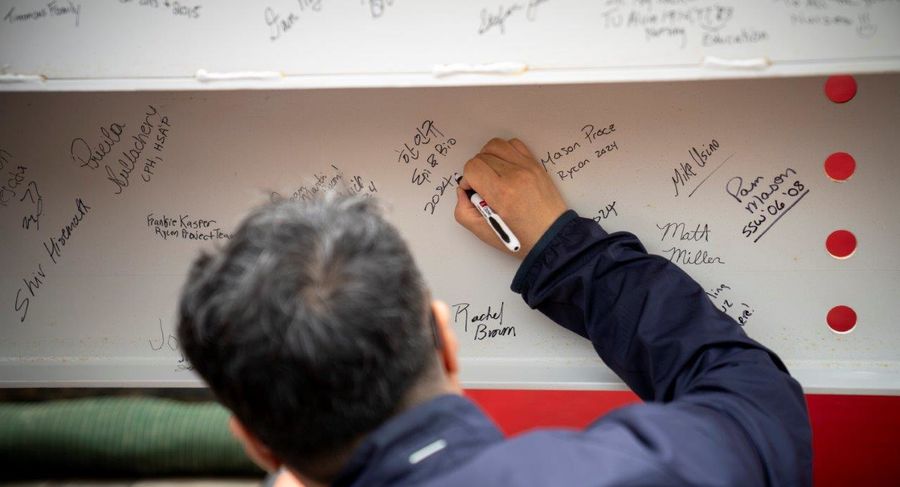 A man signs his name on a white iron beam. Other signatures are already written.