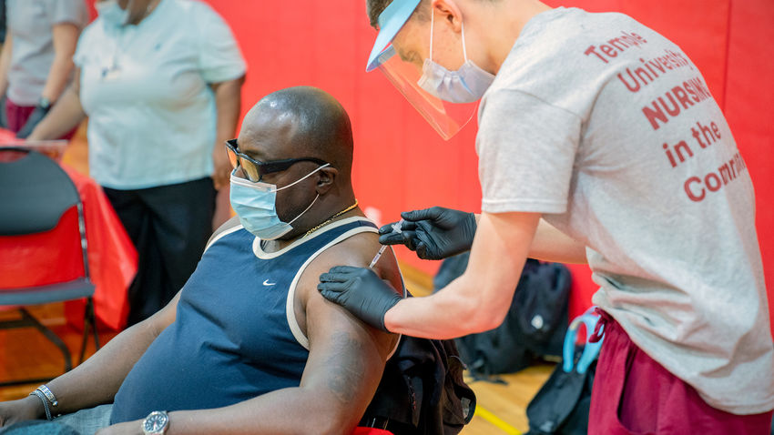 a nursing student administers a covid-19 vaccine into the arm of a seated man in a gymnasium.