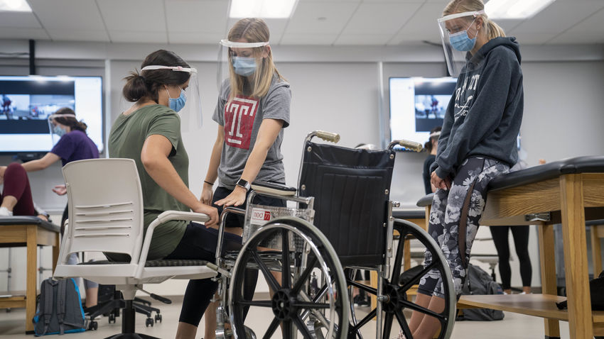 two OT students help one student move from a chair to a wheelchair