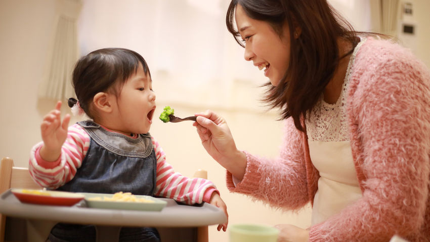 Child sitting in high chair being fed broccoli on fork by mom