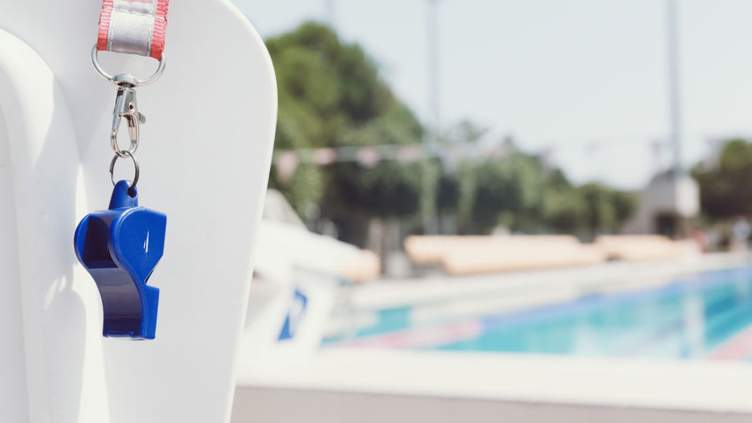 a blue whistle hangs on a lifeguard chair with a public community pool in the background