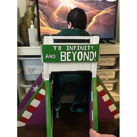 a child sits in a chair decorated as a rocket ship, with the words "to infinity and beyond" written on the chair.