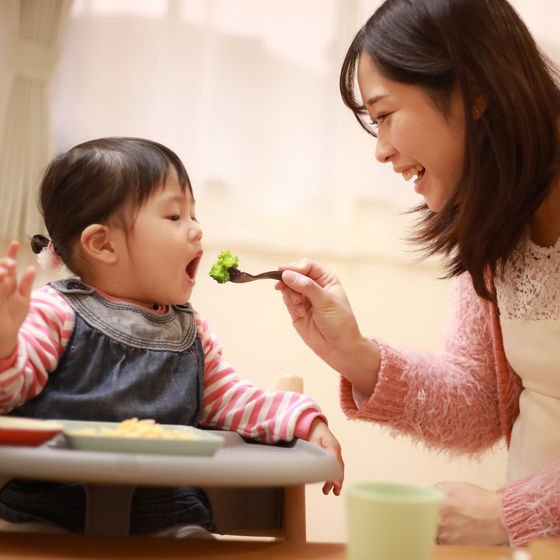 Child sitting in high chair being fed broccoli on fork by mom