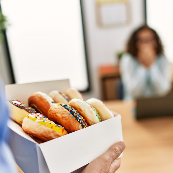 an office worker carries in a box of donuts to a conference room as a fellow coworker, out of focus, holds their hands to their mouth in surprise.