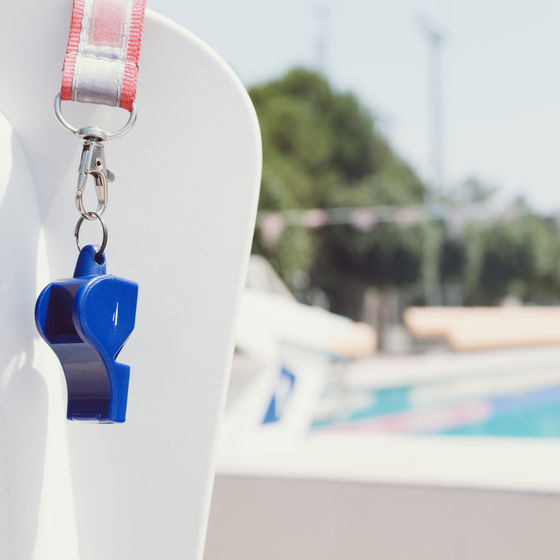 a blue whistle hangs on a lifeguard chair with a public community pool in the background